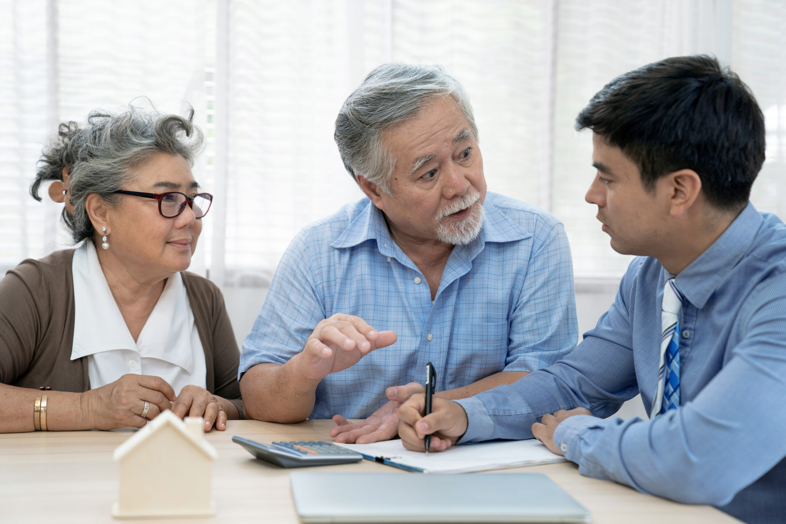 three people sitting on at a table. on the table is a calculator, paper, and a lap top computer that is closed. Two of the people are a elderly man and woman. They are speaking with a younger man . Man is dressed ina button up shirt with a tie. The purpose of this picture is to demonstrate the importance of health care planning.