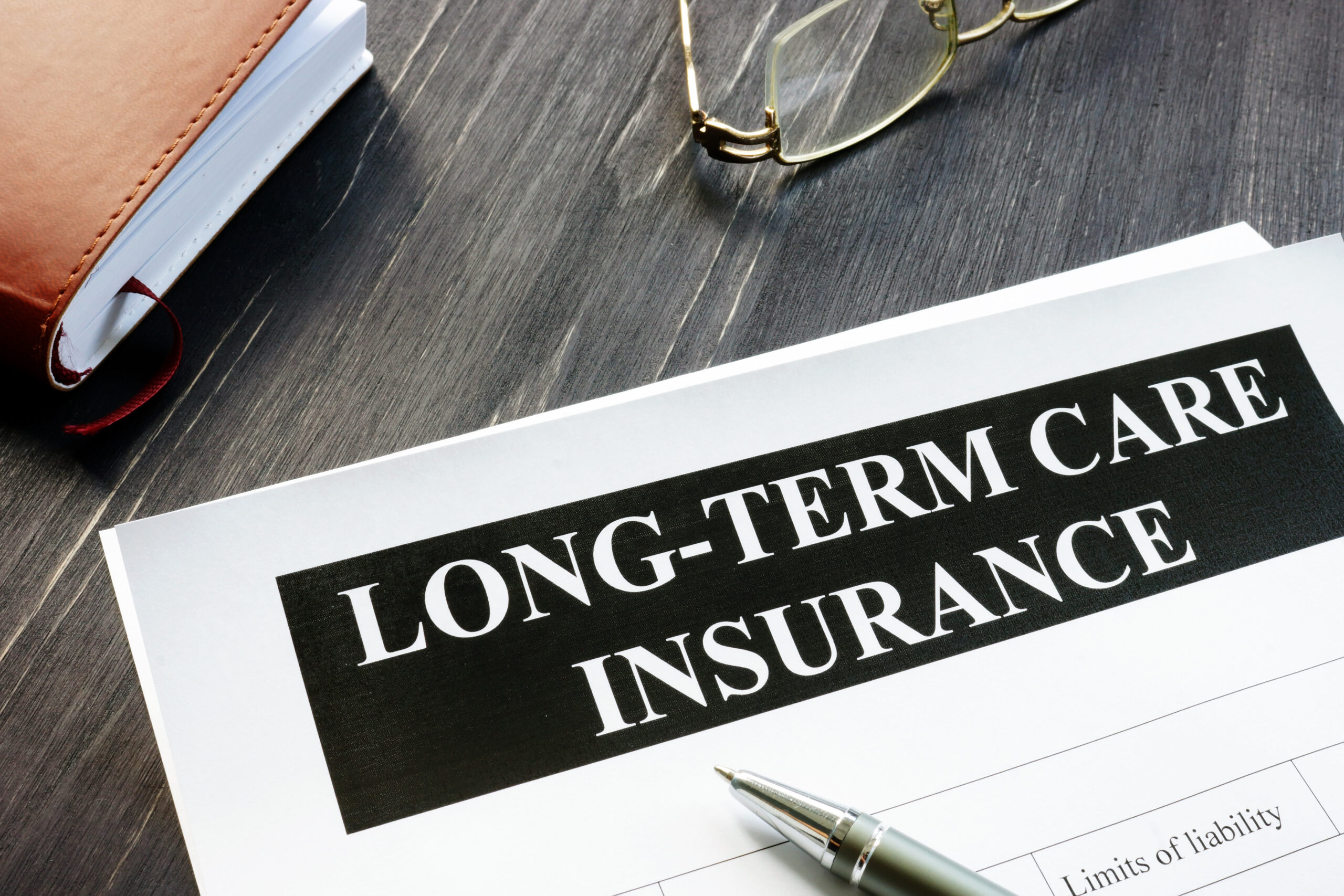 Purpose of Image: Long-Term Care Insurance. Image: background wood grained desk, eyeglasses with metal frame resting upside down on the desk, to the side of the eyeglasses in a notebook, below lays a form with the title” Long-Term Care Insurance” a silver pen rests on the form.