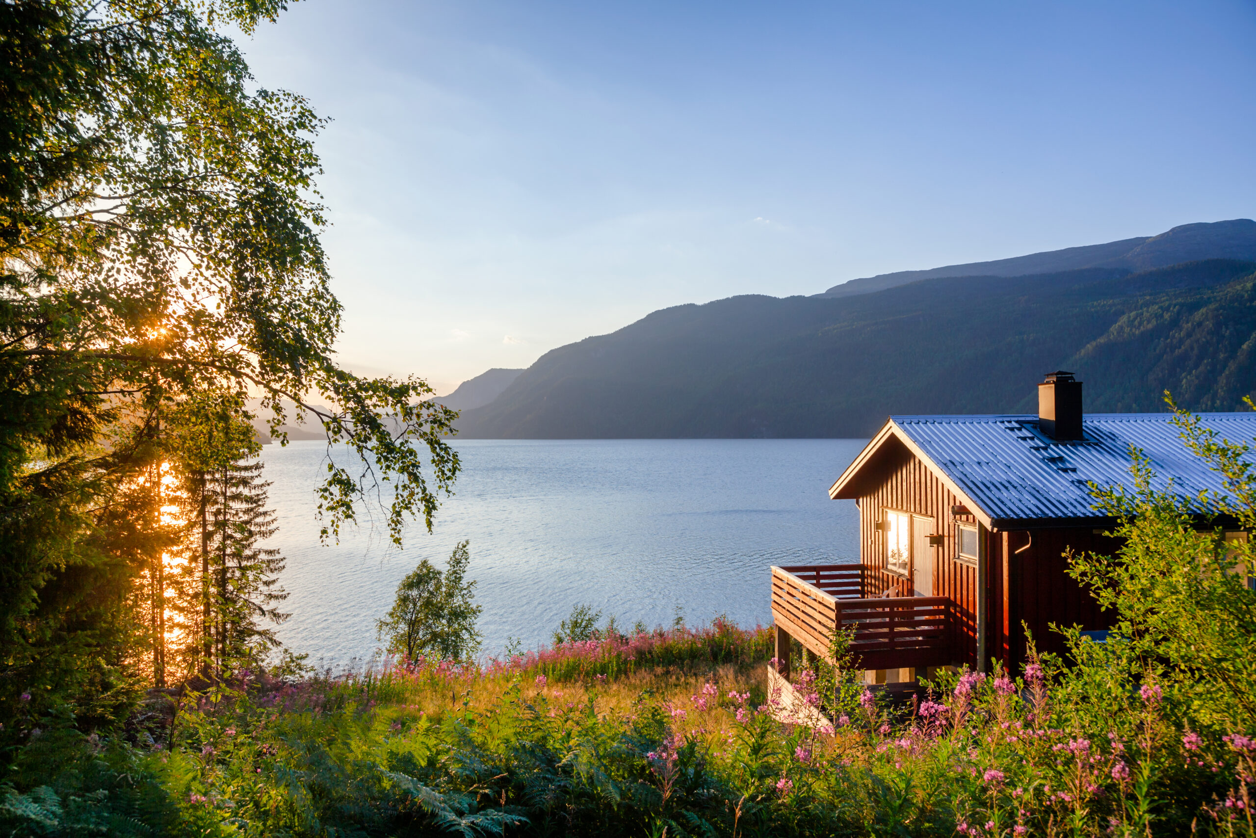 Purpose of this image: support article on learning how to include a vacation home in the estate planning. Image: wooden cabin nestled on the top on a hill surrounded by green forage. In the background is a a body of water and mountains.