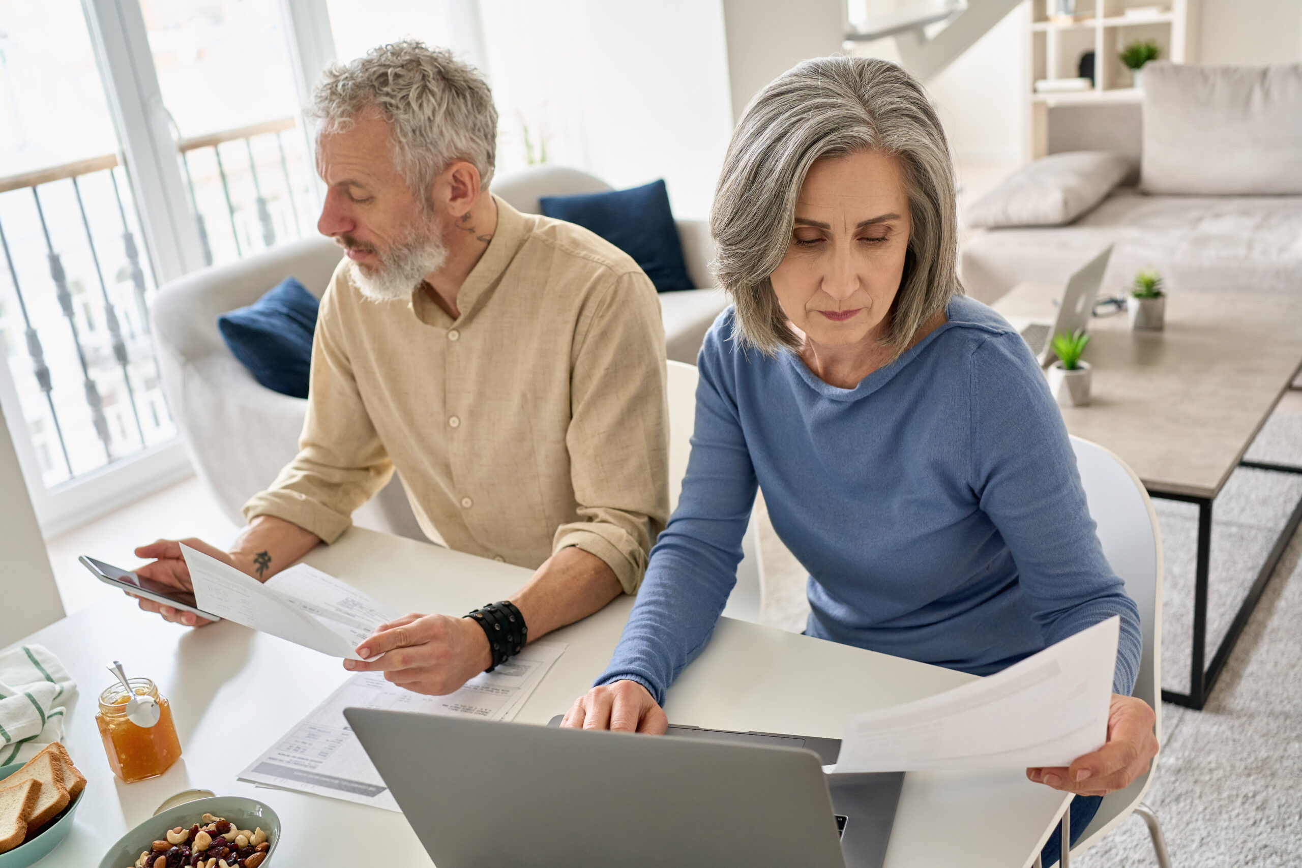 Purpose of image: Joint Accounts Image: a man an woman, siting on a white chairs with their arms resting on a white table. On the table is a computer and documents. Woman has one hand on the keyboard of the computer and looking at a document in the other. The man is holding a document in one hand and has a calculator in the other hand