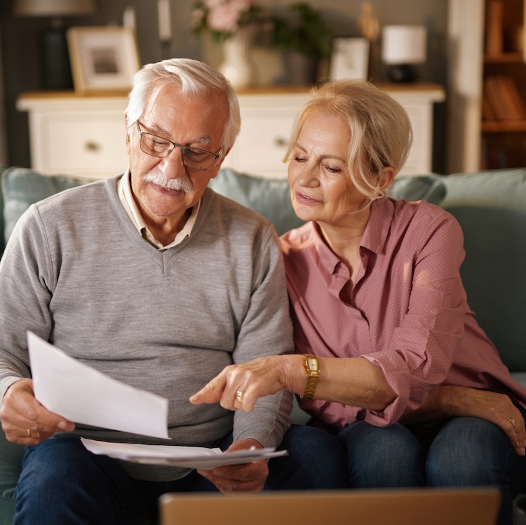 An elderly couple, sitting on a sofa, reviews paperwork and a laptop. The man holds the documents while the woman points at one. They appear to be planning their financial future together at home.
